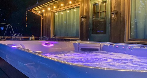 A lit hot tub with purple lights, set against a cozy wooden house at night.