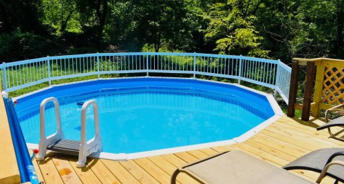A blue octagonal pool surrounded by a wooden deck and white railing, set in a lush green backyard.