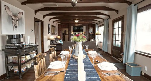 A cozy dining room with a long wooden table set for a meal, featuring rustic decor and a deer painting on the wall.