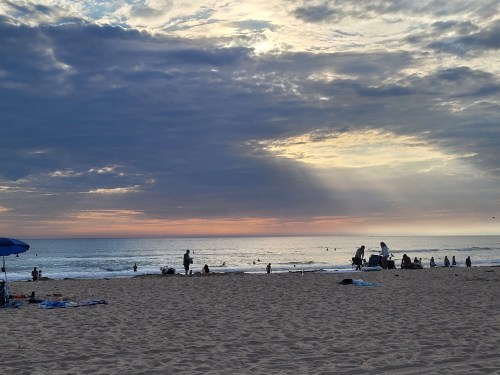 A beach scene at sunset with people enjoying the water, clouds overhead, and soft rays of sunlight breaking through.