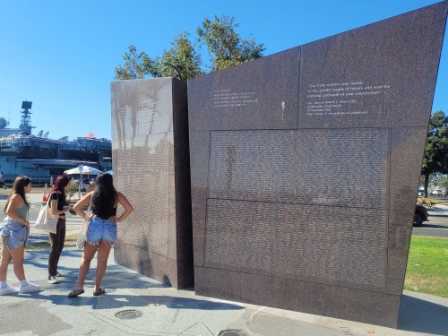 Three people stand in front of a large granite memorial with engraved text, set against a clear blue sky.