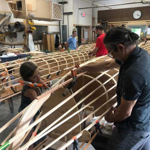 A group of people, including a child, work together on wooden boat frames in a workshop. Tools and materials are visible.