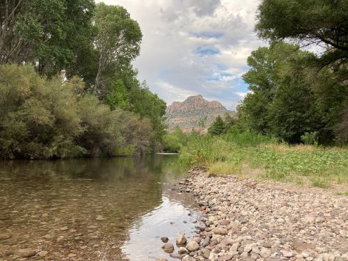 A serene riverbank with smooth stones, lush greenery, and a distant mountain under a partly cloudy sky.