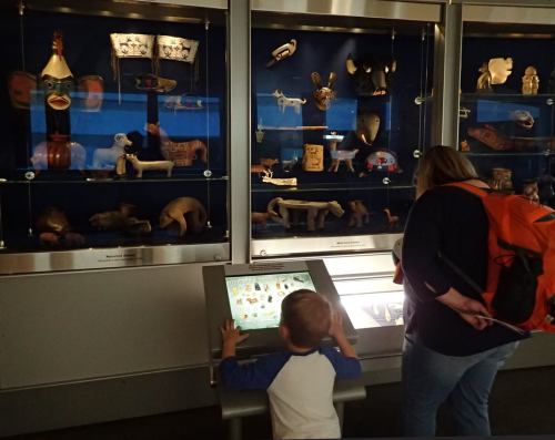 A child explores an interactive display while an adult examines artifacts in a museum exhibit.