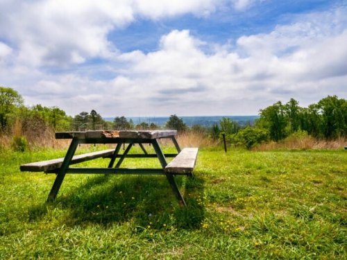 A wooden picnic table sits on green grass, overlooking a scenic view under a partly cloudy sky.