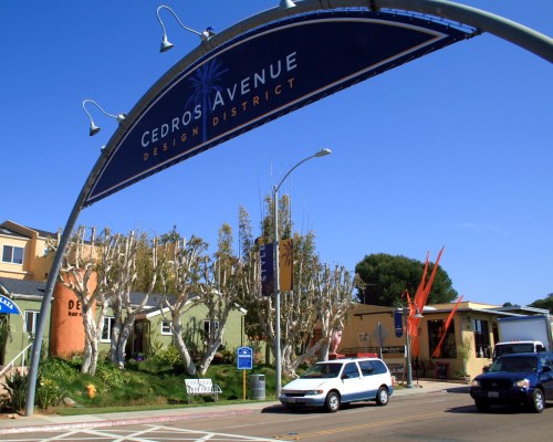 Sign for Cedros Avenue Design District, with shops and trees lining the street, and cars passing underneath.
