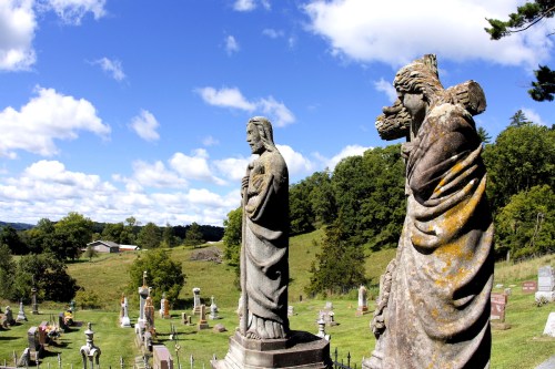 Two weathered statues of figures stand in a cemetery, overlooking a lush green landscape under a blue sky with clouds.