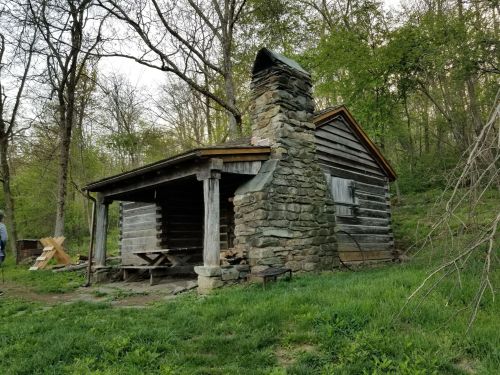 A rustic log cabin with a stone chimney, surrounded by trees and greenery.