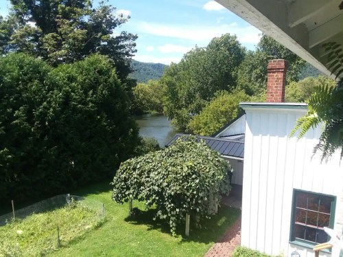 A scenic view of a river surrounded by trees, with a house and garden in the foreground under a clear blue sky.