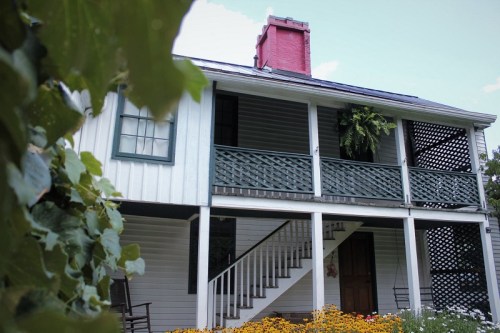 A two-story house with a red chimney, green shutters, and a porch surrounded by flowers and greenery.