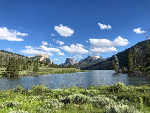 A serene landscape featuring a calm lake surrounded by mountains and lush greenery under a blue sky with fluffy clouds.