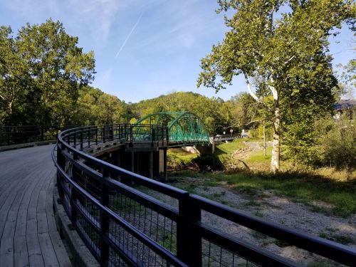 A curved wooden path leads to a green bridge over a stream, surrounded by trees and a clear blue sky.
