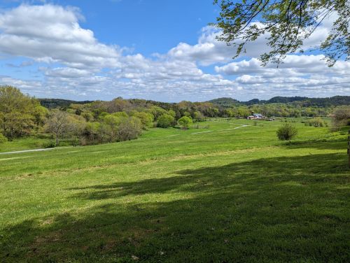 A scenic view of a green landscape with rolling hills, trees, and a partly cloudy blue sky.