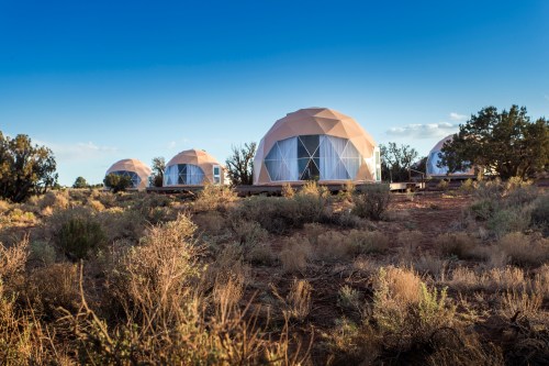 Four geodesic dome structures in a desert landscape under a clear blue sky, surrounded by sparse vegetation.