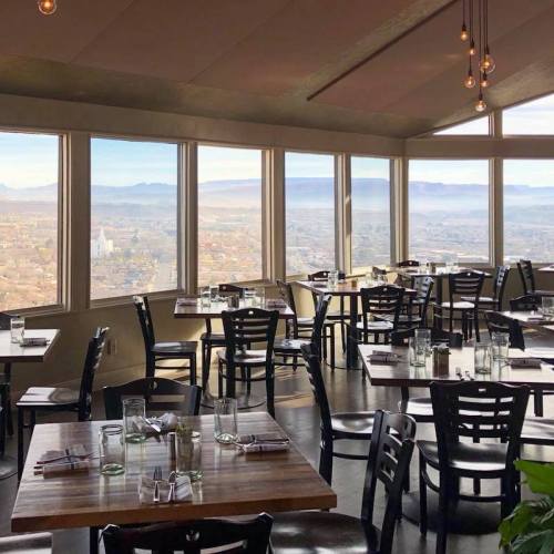 A modern restaurant with large windows offering a panoramic view of the city and mountains. Tables are set for dining.