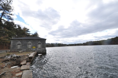 A stone building by a lake under a cloudy sky, surrounded by trees and a rocky shoreline.
