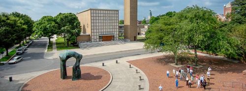 A view of a modern building with a tower, surrounded by trees and a group of people near a large sculpture.