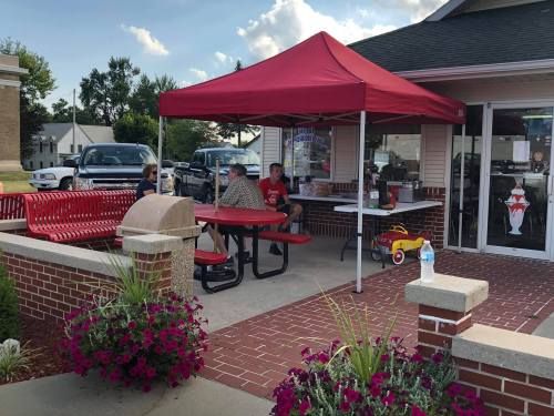 A red tent covers a picnic area outside a building, with people sitting at a table and flowers in the foreground.