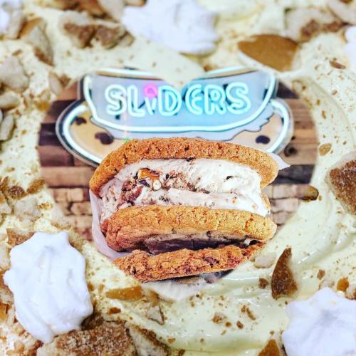 A close-up of a cookie slider filled with ice cream, surrounded by whipped cream and crumbled toppings.
