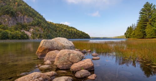 A serene lake scene with rocky shorelines, lush green trees, and calm waters under a clear blue sky.