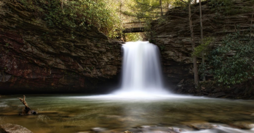 A serene waterfall cascades over rocky cliffs into a clear pool, surrounded by lush greenery and trees.