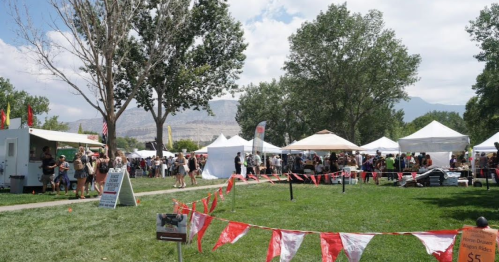 A lively outdoor festival with food stalls and tents, surrounded by trees and mountains under a blue sky.