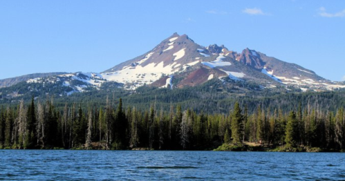 A scenic view of a snow-capped mountain rising above a forested landscape, reflected in a calm lake.