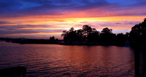 A vibrant sunset over a calm lake, with colorful skies and silhouettes of trees along the shoreline.