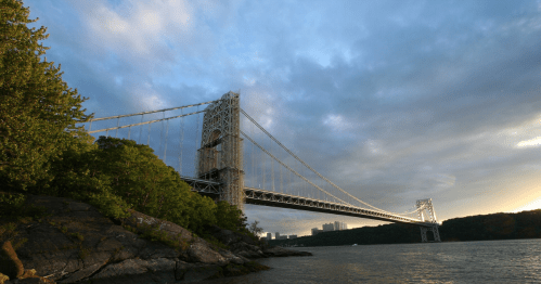 The George Washington Bridge spans a river, surrounded by greenery and a cloudy sky at sunset.
