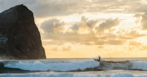 A surfer rides a wave at sunset, with a rocky cliff and clouds in the background.