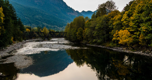 A serene river reflects autumn trees and mountains under a clear sky, with a bridge crossing the water.