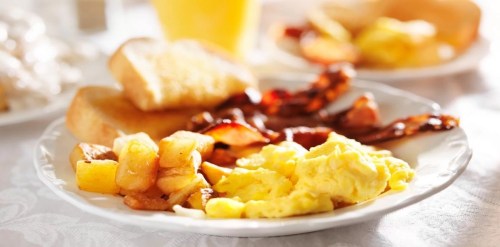 A plate of breakfast featuring scrambled eggs, bacon, toast, and diced potatoes, with a glass of juice in the background.