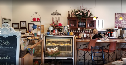 Cozy café interior with a display case, counter, and floral decor; a customer sits at the bar enjoying their drink.