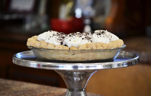 A delicious pie on a silver stand, topped with whipped cream and chocolate shavings, set against a blurred background.