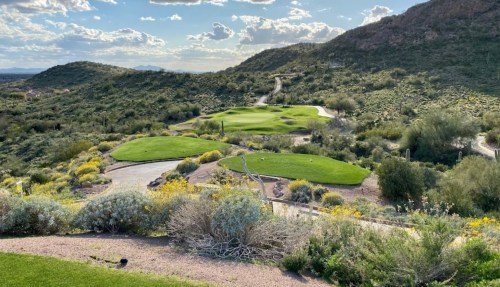 A scenic view of a golf course surrounded by lush greenery and rolling hills under a partly cloudy sky.