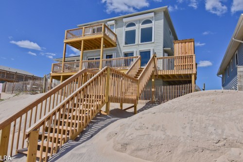 A beach house with wooden decks and stairs leading down to sandy dunes under a blue sky with scattered clouds.
