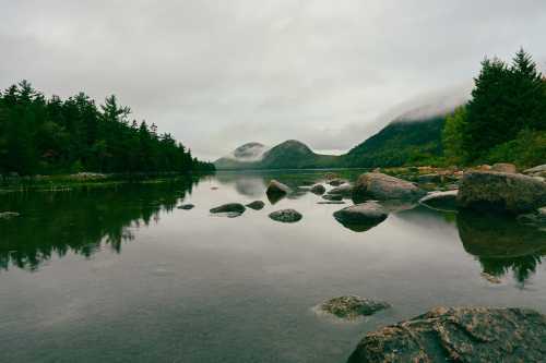 A serene lake surrounded by trees and mountains, with rocks in the foreground and misty clouds overhead.