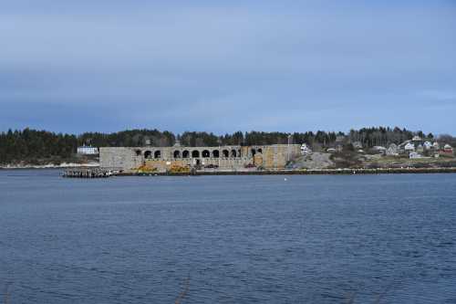 A stone structure by the water, surrounded by trees and houses, under a cloudy sky.