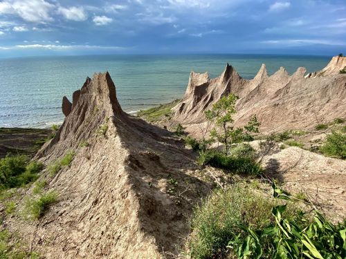 A rugged coastline with sharp, rocky formations and lush greenery, overlooking a calm sea under a cloudy sky.