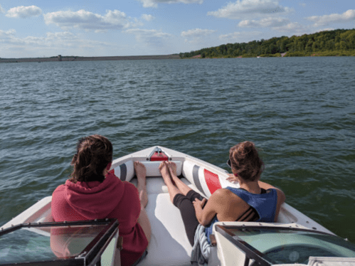 Two people relax on the bow of a boat, facing a calm lake with trees and a dam in the background under a blue sky.