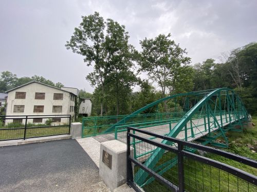 A green metal bridge spans a small river, with a wooded area and a building in the background under a cloudy sky.