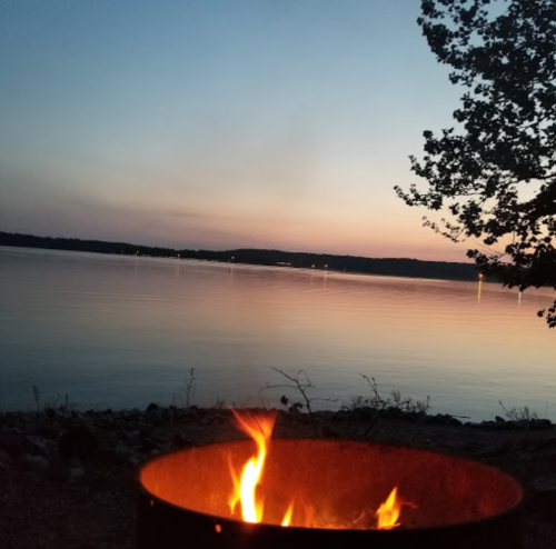 A campfire burns in a metal ring by a calm lake at sunset, with trees silhouetted against a colorful sky.