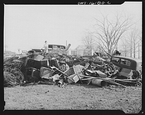 A large pile of scrap metal and debris, including old cars, in a barren landscape with bare trees in the background.