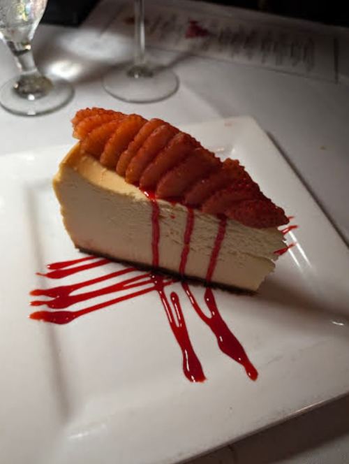 A slice of cheesecake topped with fresh strawberries and drizzled with raspberry sauce on a white plate.