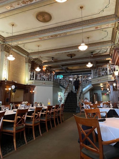 Elegant restaurant interior with wooden tables, white tablecloths, and a grand staircase leading to an upper level.