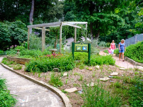 Two women walk through a lush garden with a wooden structure and colorful flowers along a winding path.