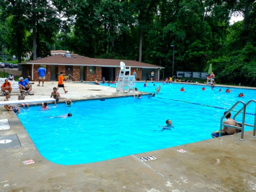 A community pool scene with people swimming, lounging, and enjoying the water on a sunny day surrounded by trees.