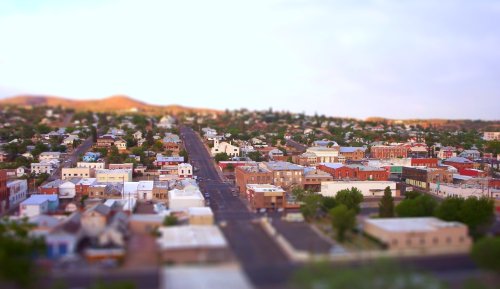 Aerial view of a small town with colorful buildings, streets, and hills in the background under a soft, bright sky.