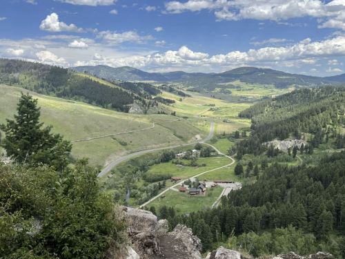 A scenic view of rolling hills, a winding road, and a small farm nestled in a lush green valley under a blue sky.