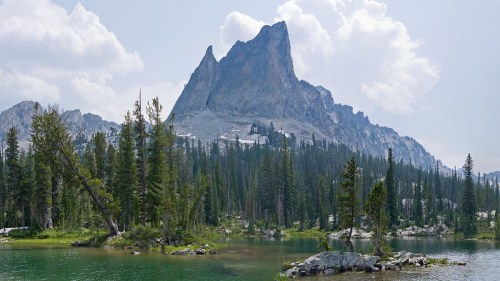 A majestic mountain rises above a serene lake, surrounded by lush green trees and a cloudy sky.